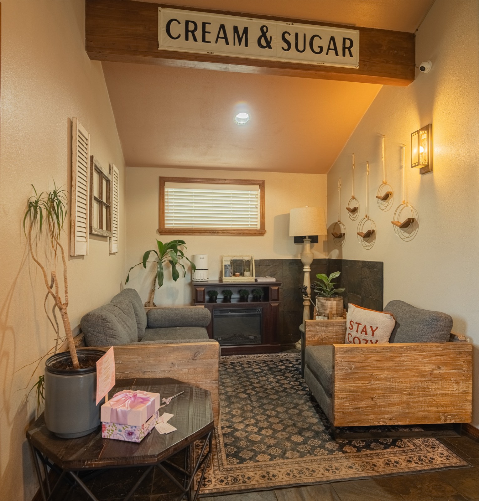 The lounge nook inside Cozy Coffee under the Cream and Sugar sign, with a fireplace, soft chairs, and a Stay Cozy pillow